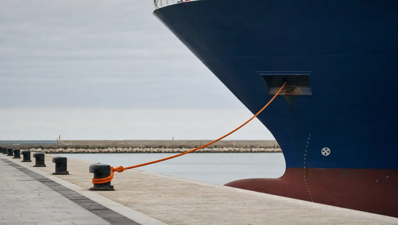 The towering navy bow of a cargo ship held against a stone harbour quay by a single taut orange mooring line to one bollard while the other bollards stand empty, illustrating an unmanaged single point of failure.